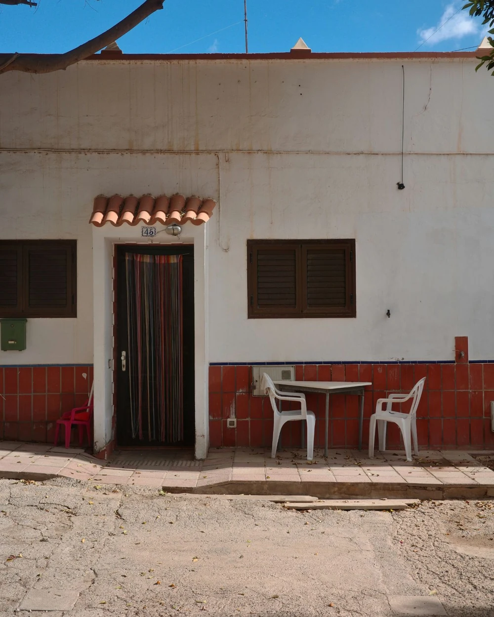 Fachada blanca de una casa con una cortina de cintas en la puerta y dos sillas afuera
